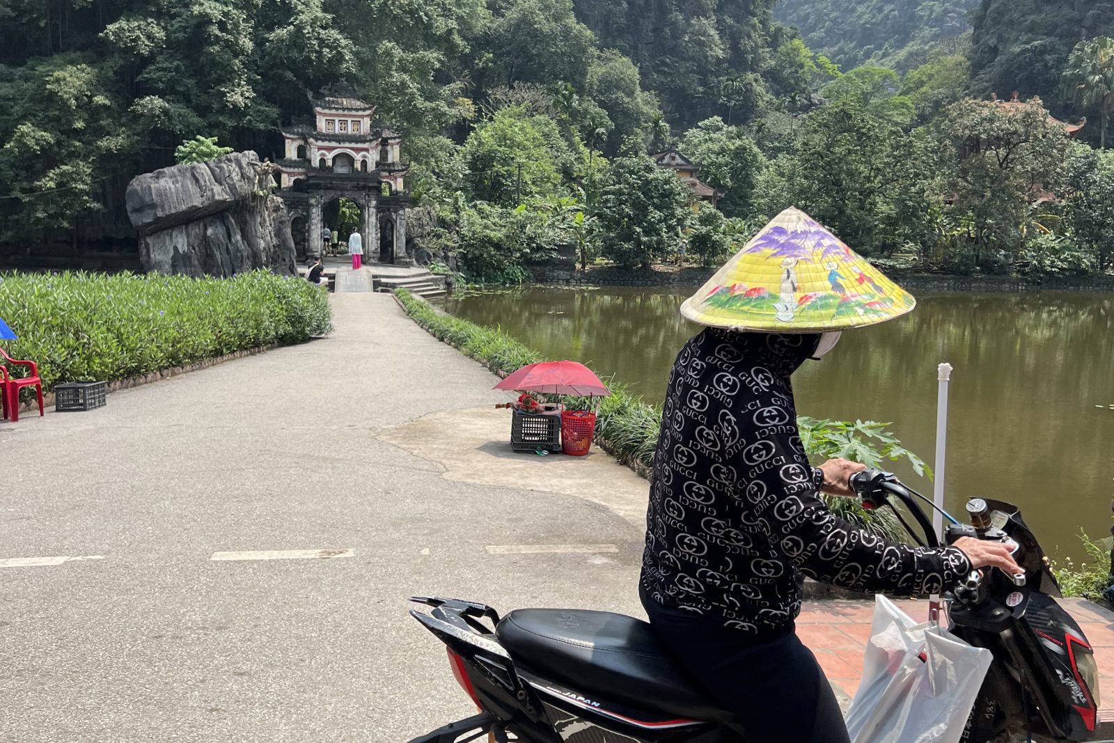 Mulher de chapéu cônico parada de moto diante do portal do pagode e do lago, em Ninh Bình, Vietnã