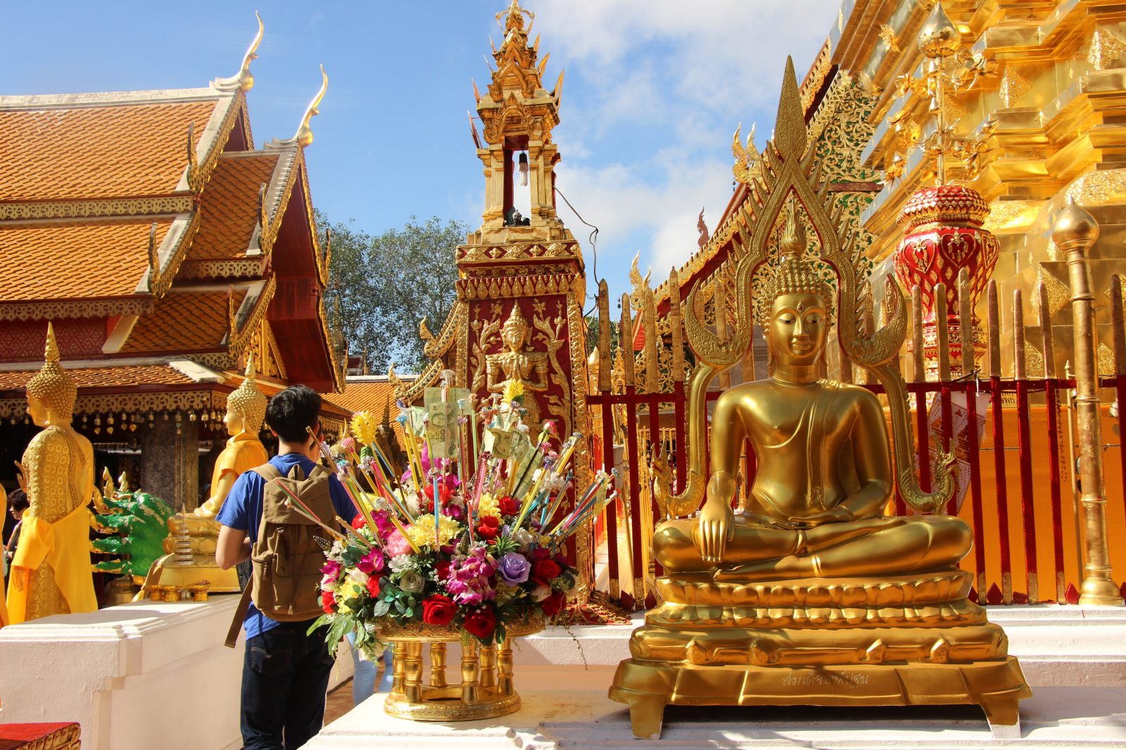 Visitante de mochila, de costas, diante de um arranjo de flores e de um Buda dourado em um templo de Chiang Mai