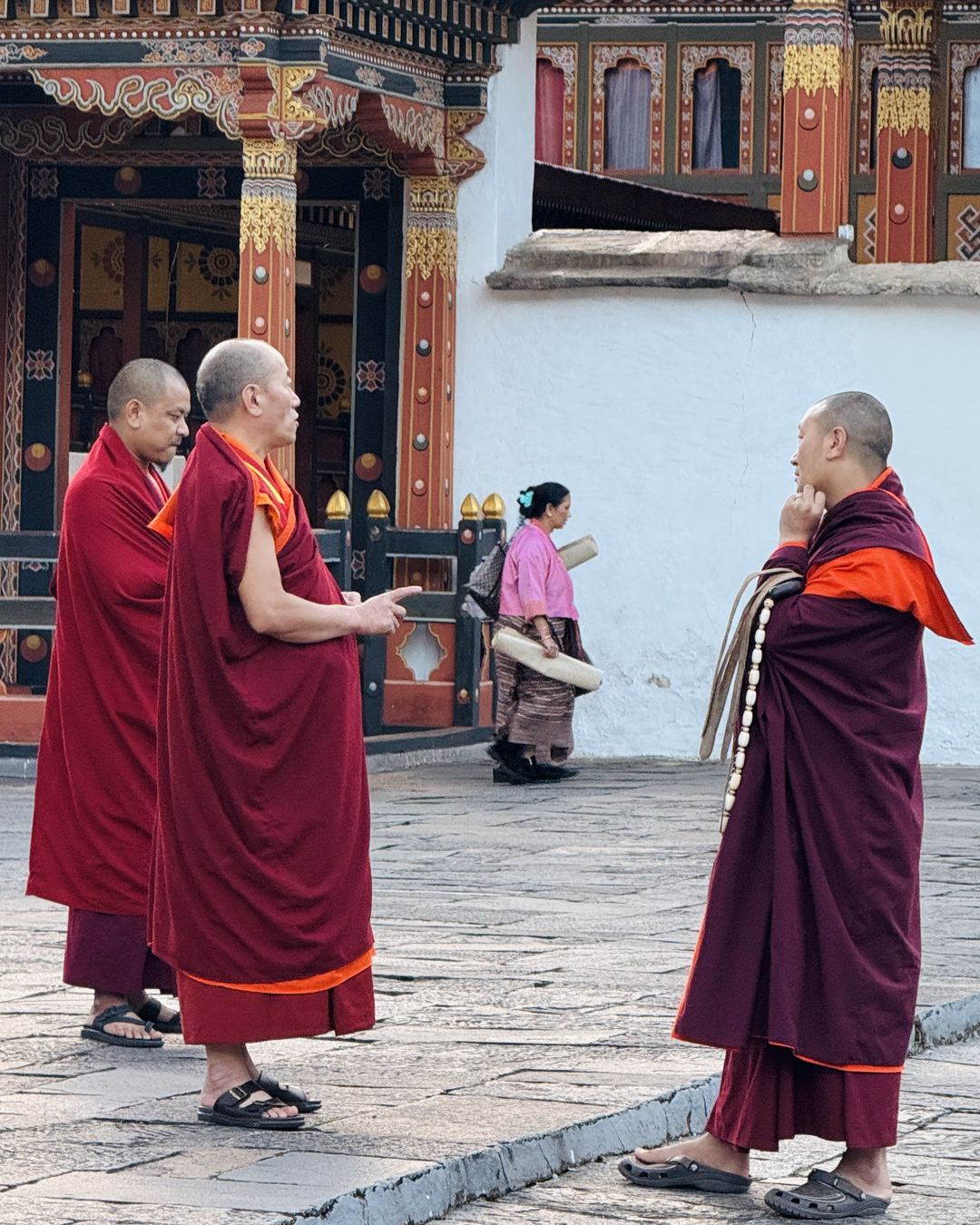 Três monges de manto vinho conversando no pátio de um dzong butanês, diante das colunas pintadas